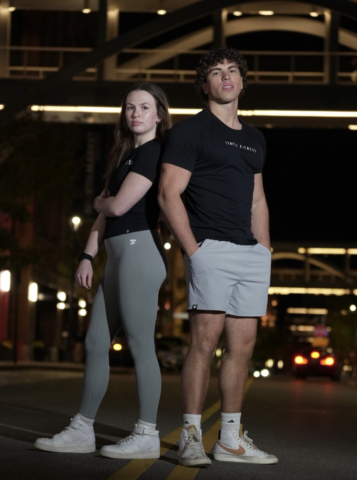 Temple Fitness duo standing back to back on a city street at night